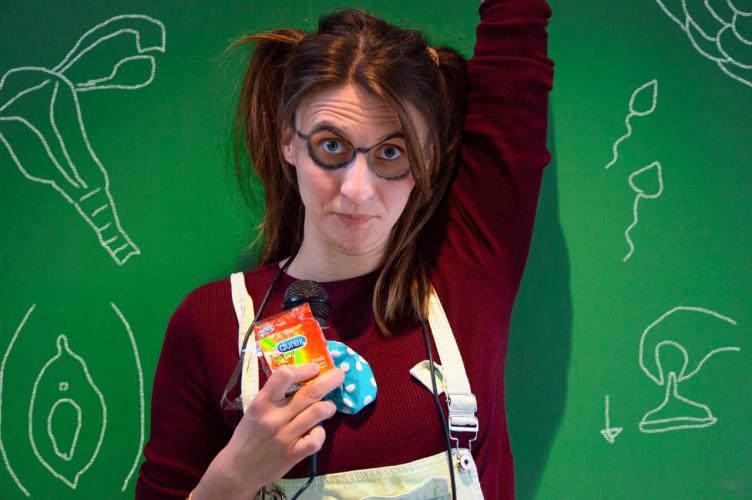 Performer Holly Delefortrie has drawn on glasses and her hair is in bunches. In one hand she holds condoms, balloons and a microphone, the other hand is raised as if to ask a question.  Holly is in front of green-chalkboard with pictures of uterus, vulva and sperm behind her.