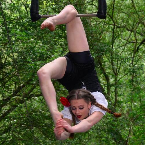 Woman hanging from a trapeze from one leg, with trees in the background
