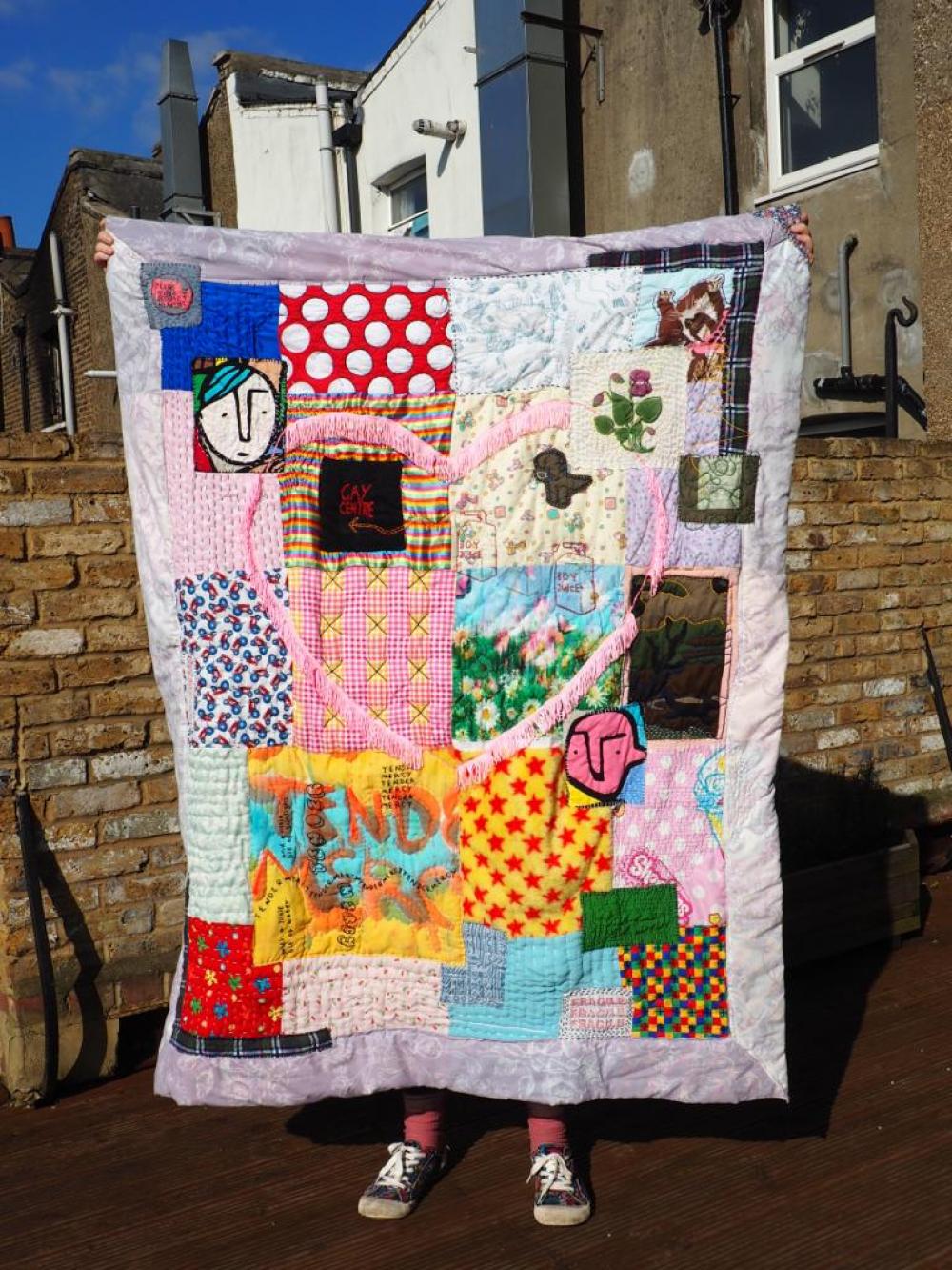 Colourful quilt artwork held up against blue sky and houses.
