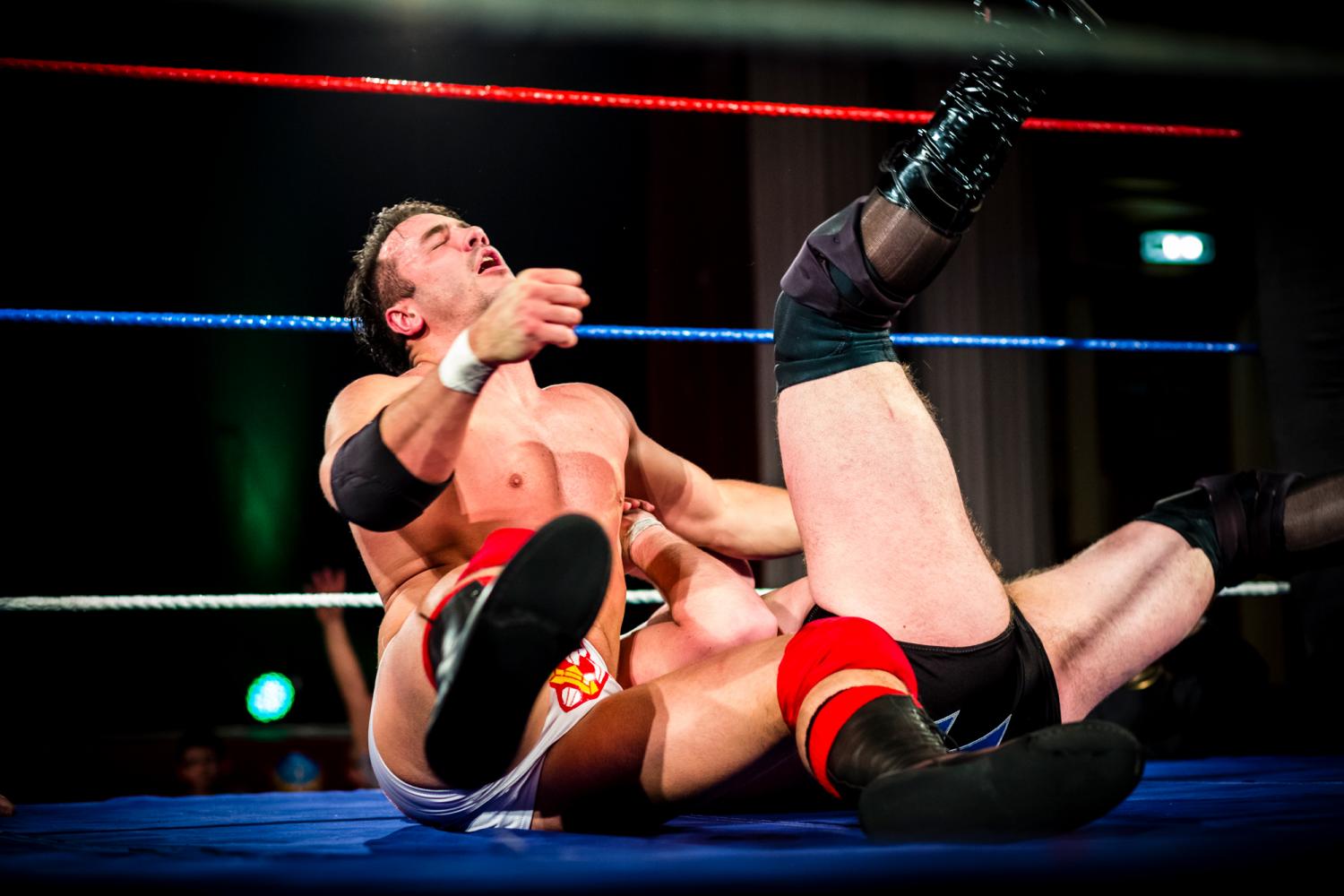 A male wrestler sits exhausted in the ring, next to another more obscured wrestler with his legs in the air.