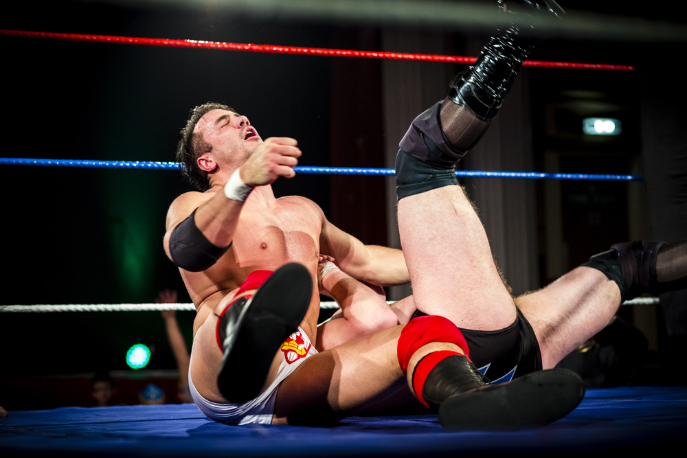 A male wrestler sits exhausted in the ring, next to another more obscured wrestler with his legs in the air.