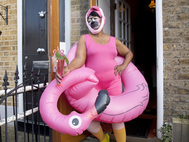 An older woman of Afro-Caribbean heritage stands in her doorway looking fierce dressed as a flamingo and in a hot pink leotard.