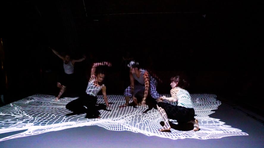 4 dancers moving energetically in a dark studio with a grid of white light projected over them onto the floor
