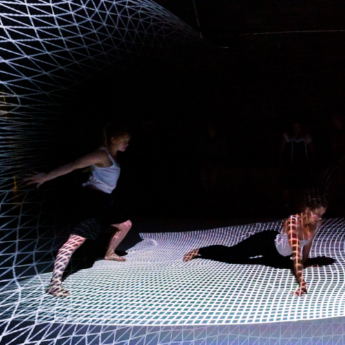 2 dancers moving energetically in a dark studio with a grid of white light projected over them onto the floor