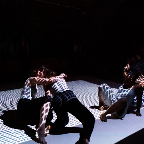3 dancers moving energetically in a dark studio with a grid of white light projected over them onto the floor