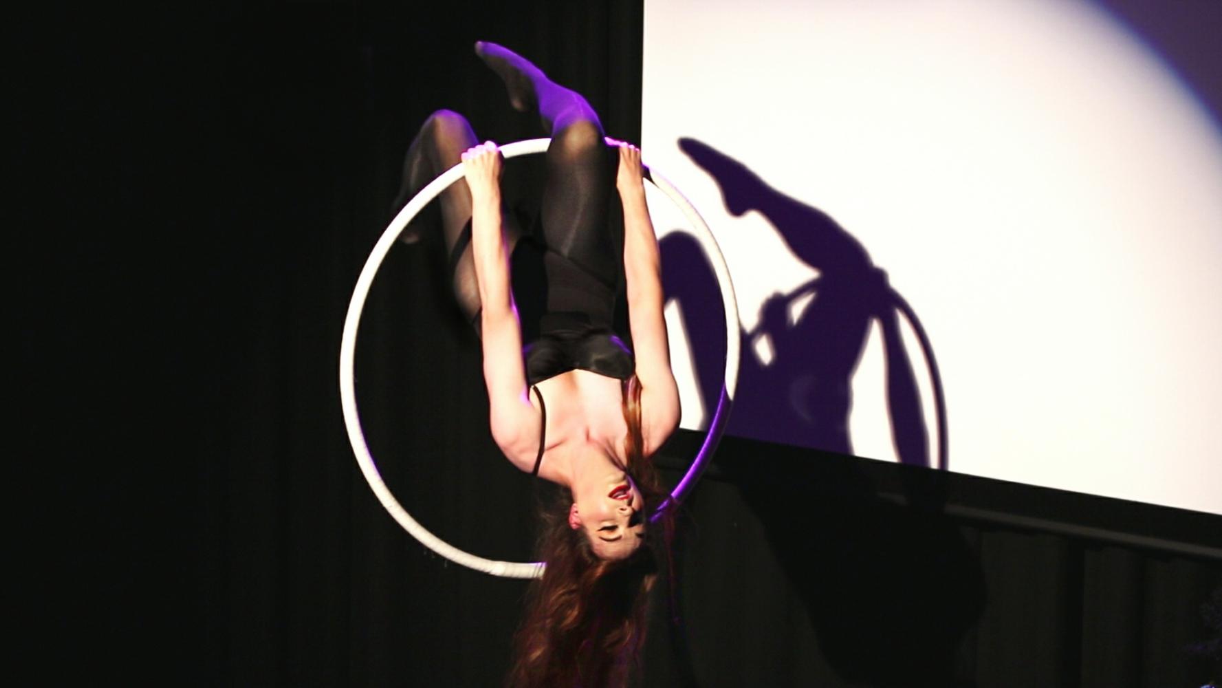 A woman in shiny black leotard & tights hangs upside down from her knee in an aerial hoop; her long red/brown hair hangs down