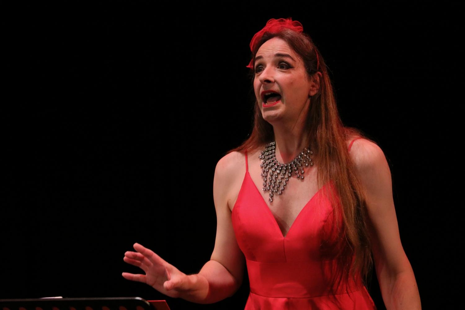 A woman with long brown hair stands in a red evening dress, behind a music stand. Her mouth is open; she is singing.