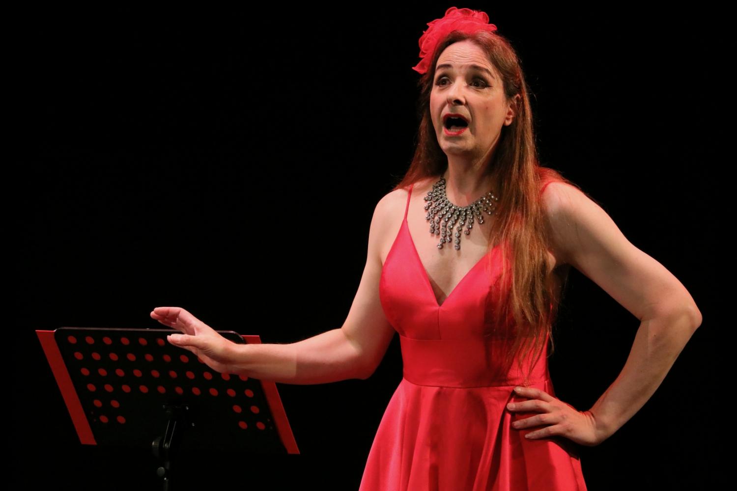 A woman with long brown hair stands in a red evening dress, before a music stand. Her mouth is open; she is singing.