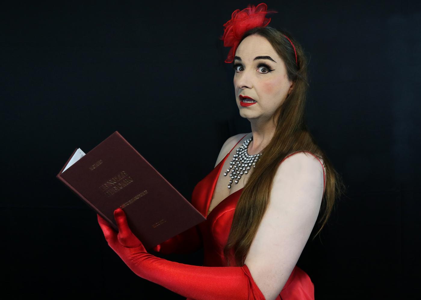 A woman with long brown hair, in a red evening dress & opera gloves is holding a music score - she looks to camera worriedly.