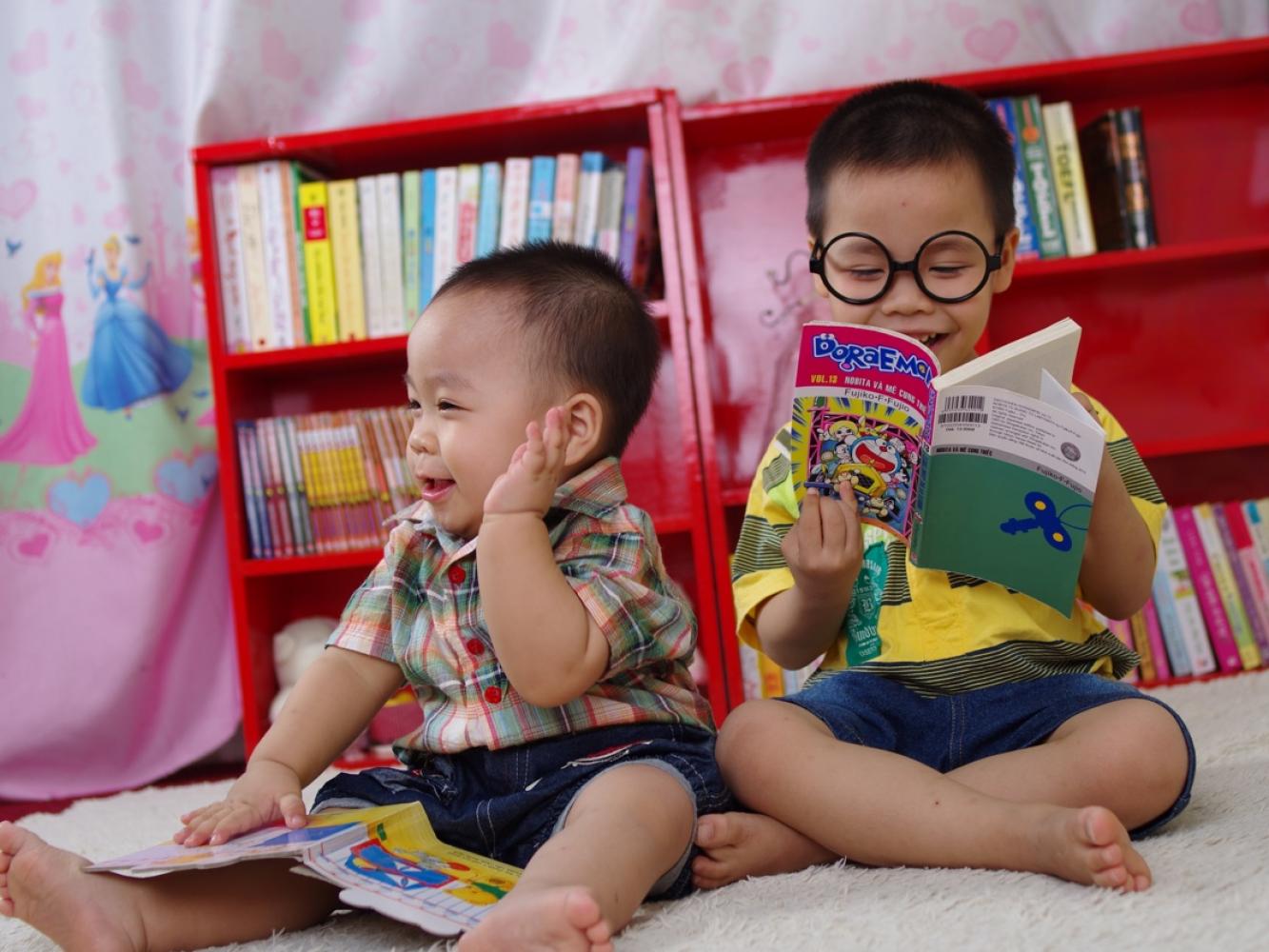 Two youngsters sit on a carpet they are sounded by colour and books.  They are both joyful with books in their hands.