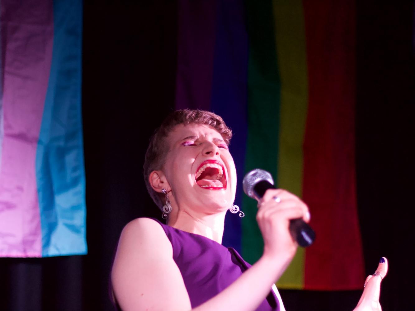 A performer is centre open mouthed singing an impressive note with a mic in their hand.  There are pride flags behind them.