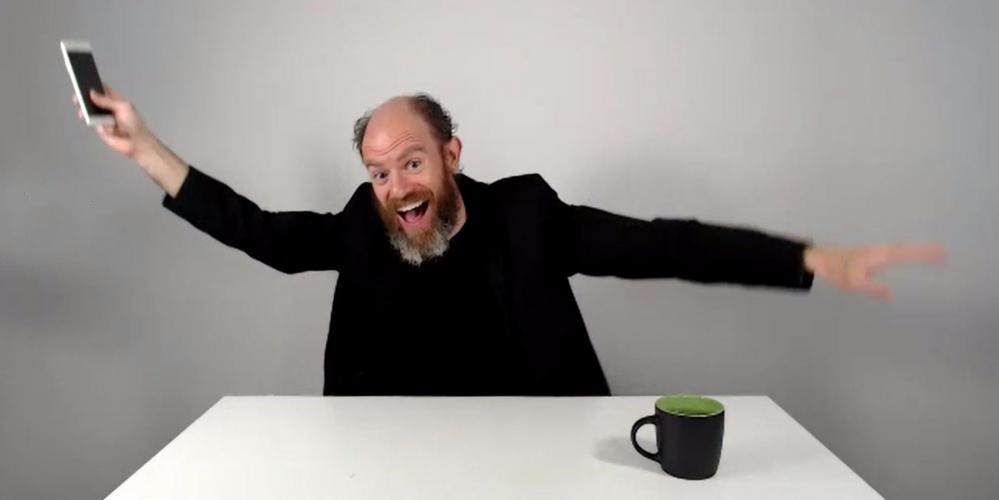 A white male, balding up top, brown hair on the sides, and a full beard, graying at the bottom, looks at the camera with his mouth open. He is dressed in all black, with a tight-fitting black jacket. Both of his arms are outstretched and in his left hand he holds an iPhone. The background is all white, as is a table in front of him with a black mug which is green inside to his right.