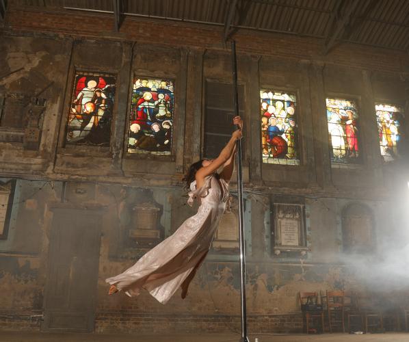 A woman flying around a pole in a long dress inside a chapel with sun pouring through the stained-glassed windows.