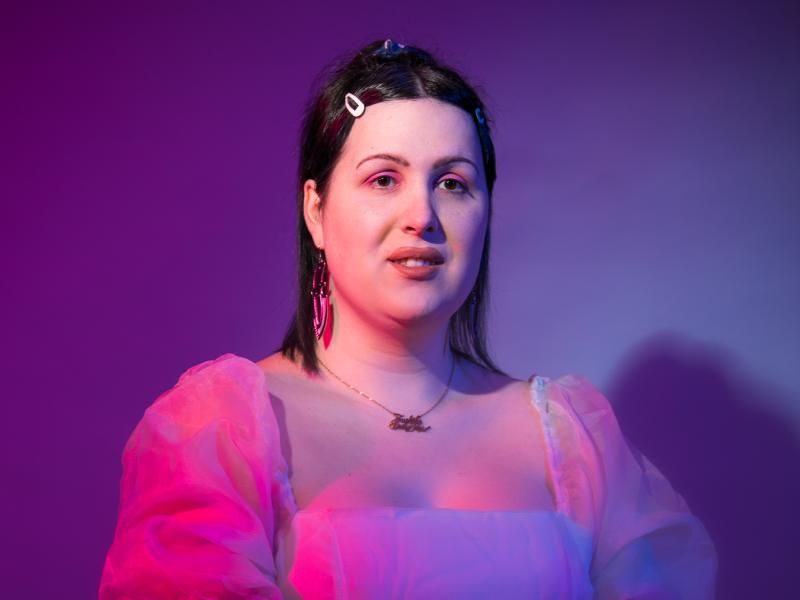 an image of Jen Ives sitting with purple light on her face, she is wearing a puffy pink dress