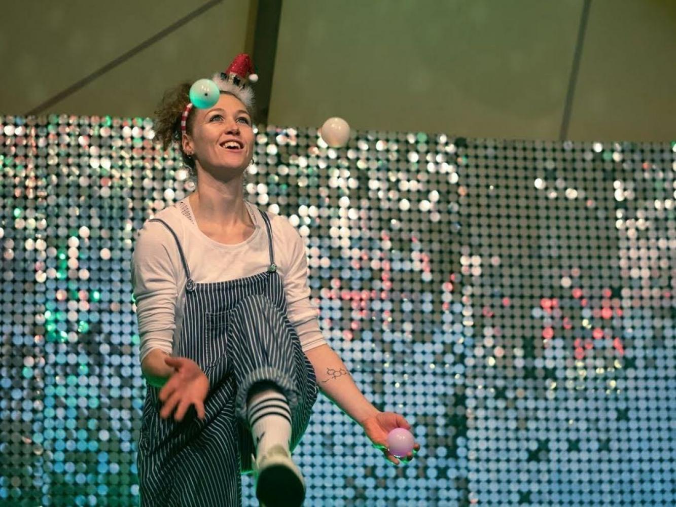 A dancer does a superhero pose in profile - one arm outstretched and a leg lifted behind her, in front of a colourful stall