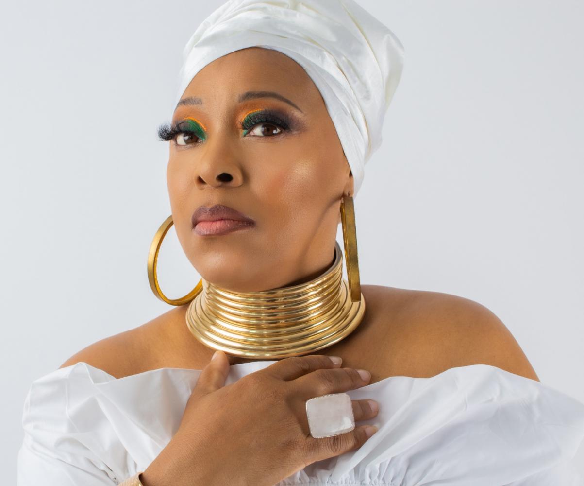 Sheba Montserrat in a white headwrap, gold choker and large hoop earrings.