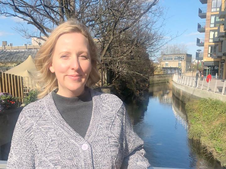 Becky, a white woman with blonde hair, is smiling with the River Wandle behind her. The river has trees on one side and a path with flats on the other. It is a sunny winters day with a blue sky.