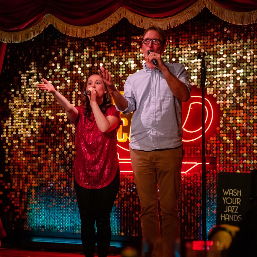 Two performers, a short female and a tall male, holding hand held microphones with hands out toward the audience.