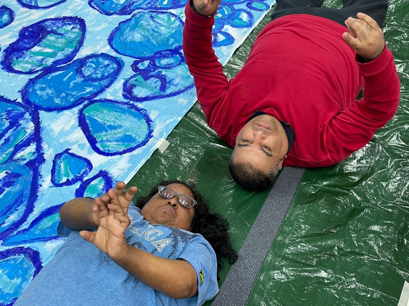 Close up of two artists lying down on the floor with their hands stretched upwards, next to colourful blue artwork