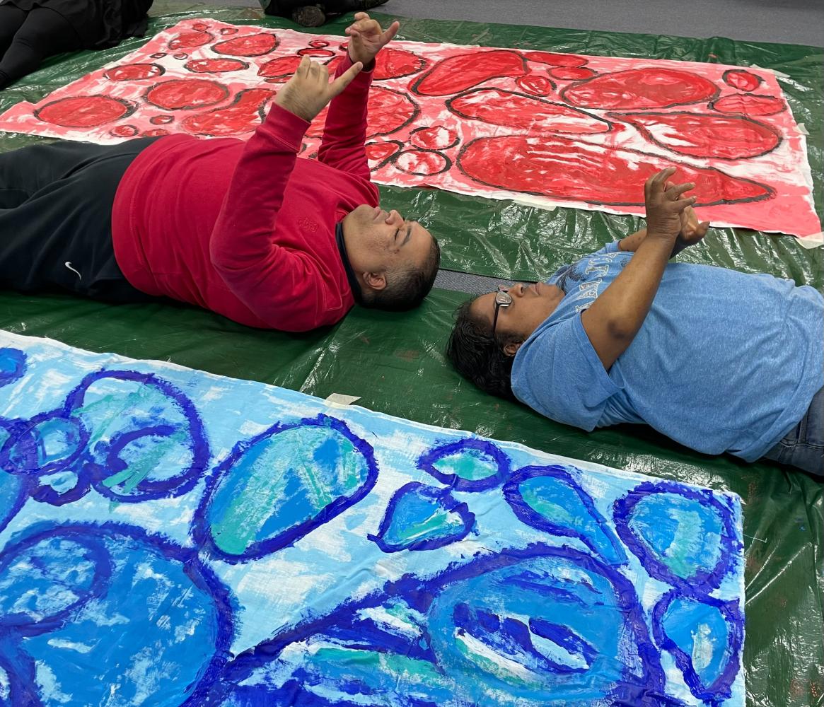 Two artists lie on the ground next to red and blue colourful artwork, reflecting their red and blue tops