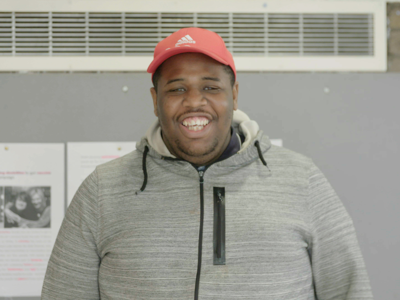 A man in a red hat and grey hoodie is looking at the camera and smiling. It is a close up shot.