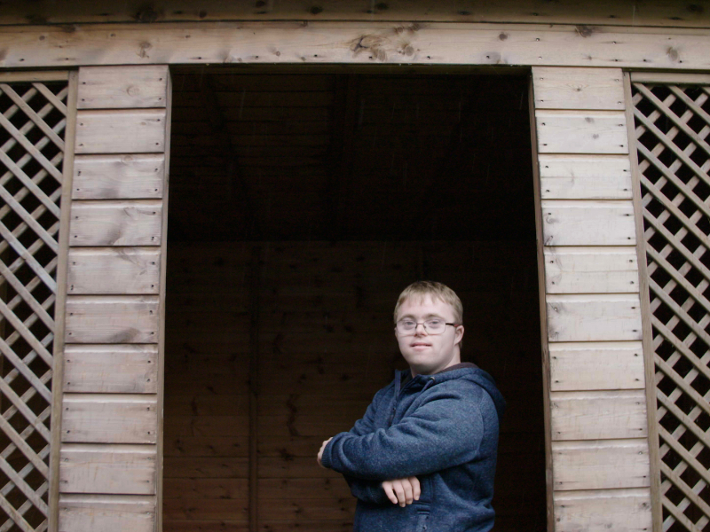 A man with Downs Syndrome stood in the doorway of a shed. He has blond hair, is wearing glasses and his arms are folded.