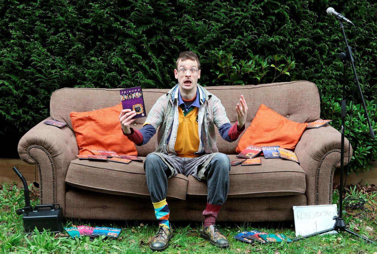 Dylan Dodds sits on a sofa in front of a hedge. He is surrounded by Friends DVD's and doesn't look happy