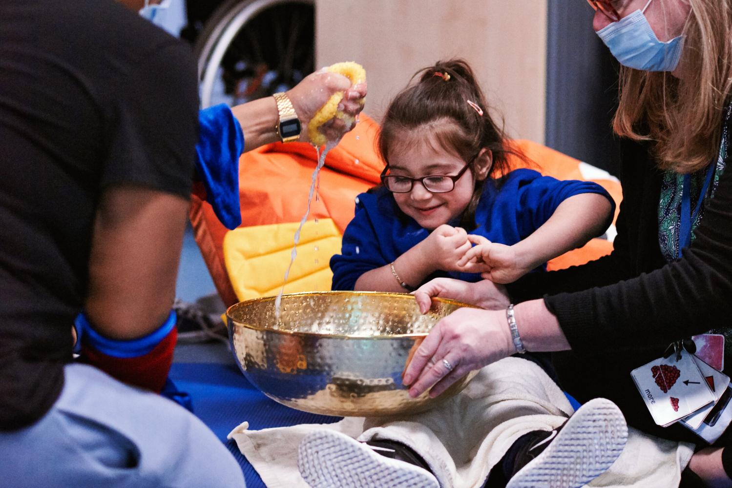 Young person sitting on a blue mat laughs as their supporting adult squeezes a sponge filled with water into a gold bowl