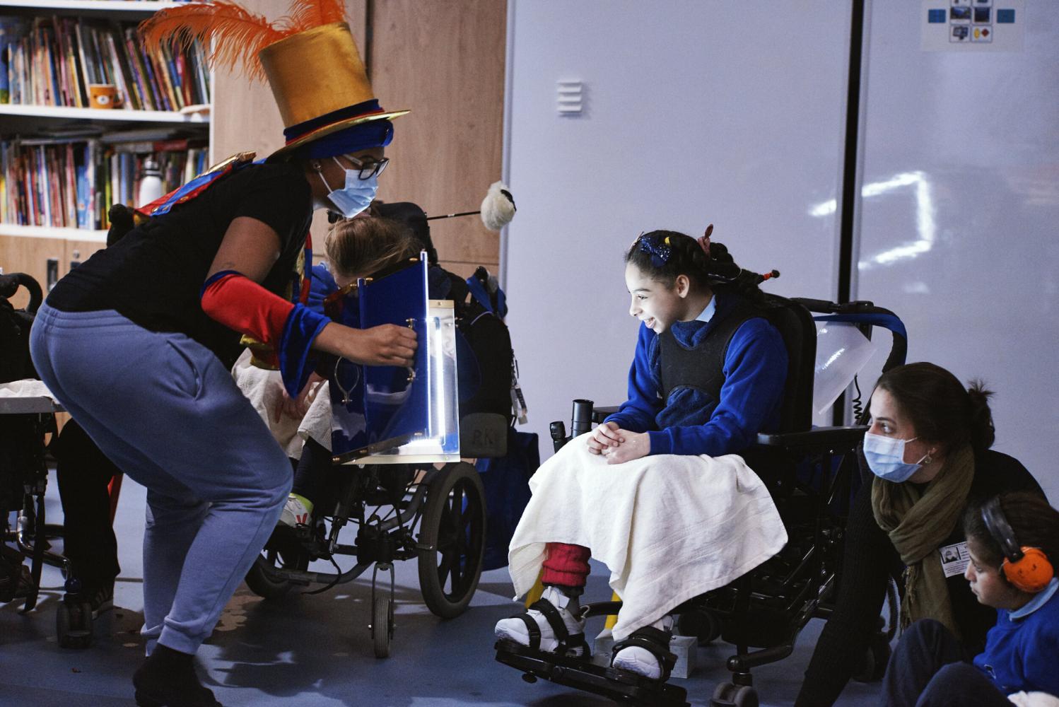Young person in a wheelchair smiles at their reflection as their supporting adult holds up a mirror