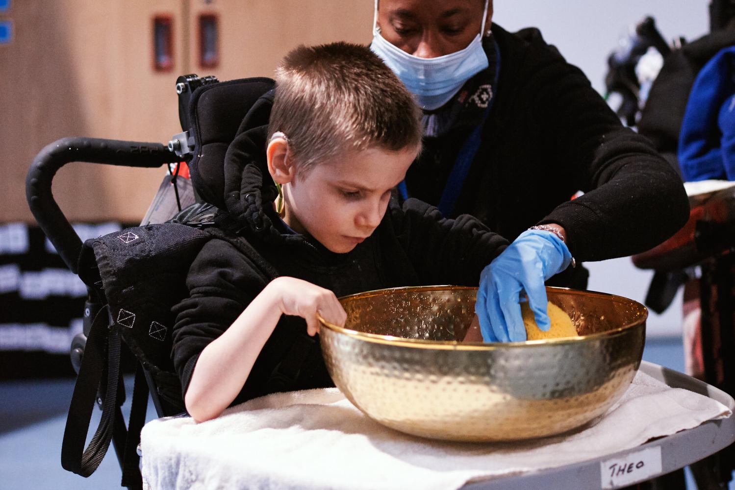 Young person in a wheelchair looks at their supporting adults hand as they squeeze a sponge in a gold bowl filled with water